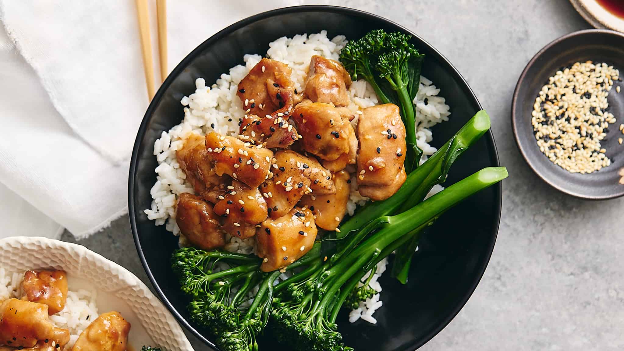 Savory teriyaki chicken with steamed rice and fresh broccoli broccolini, garnished with sesame seeds on a black plate over gray textured surface.