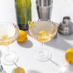 Bright lemon cocktails in elegant coupe glasses garnished with lemon twists, set on a white countertop with lemons, cocktail tools, and a green bottle in the background.