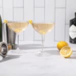 Lemon cocktails in coupe glasses with lemon garnishes, fresh lemons, a cocktail shaker, and liquor bottles on a white kitchen countertop. Perfect for summer drinks and cocktail recipes.