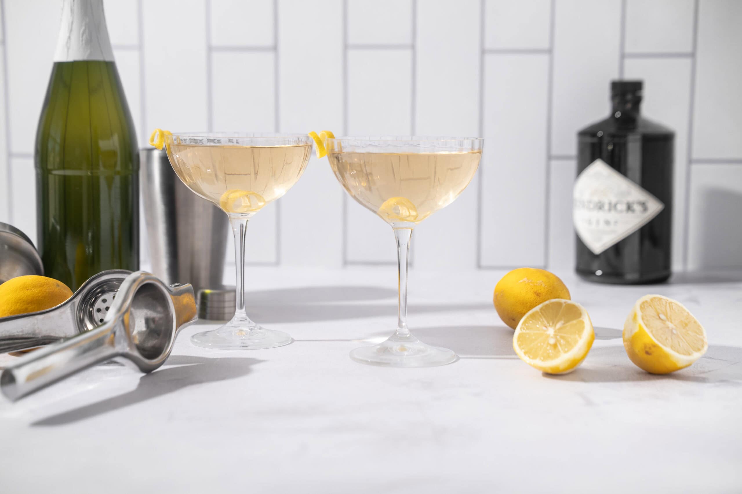 Lemon cocktails in coupe glasses with lemon garnishes, fresh lemons, a cocktail shaker, and liquor bottles on a white kitchen countertop. Perfect for summer drinks and cocktail recipes.