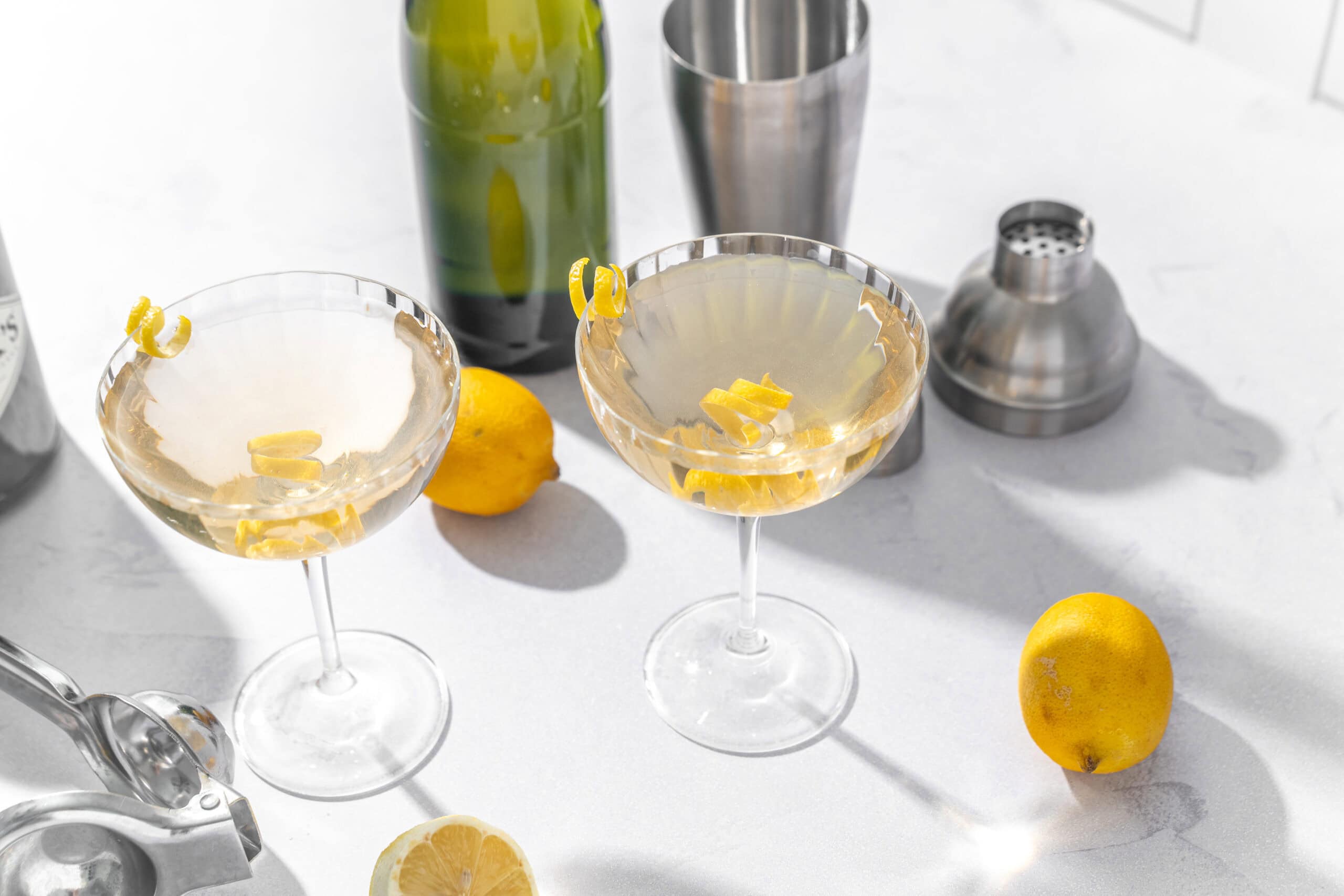 Bright lemon cocktails in elegant coupe glasses garnished with lemon twists, set on a white countertop with lemons, cocktail tools, and a green bottle in the background.