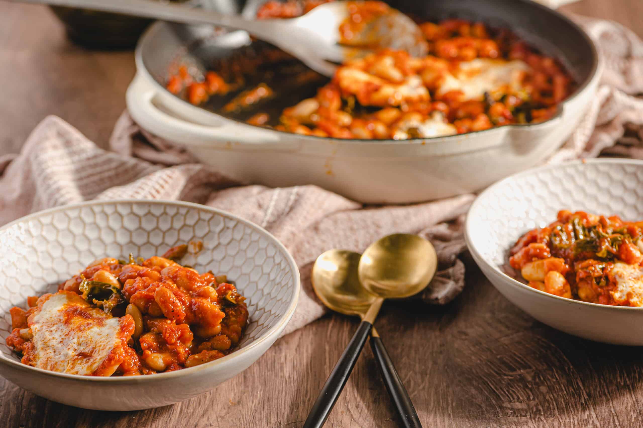 Creamy baked bean casserole with melted cheese and herbs served in decorative bowls on rustic wooden table. Perfect comfort food dish for family dinner or outdoor gathering.