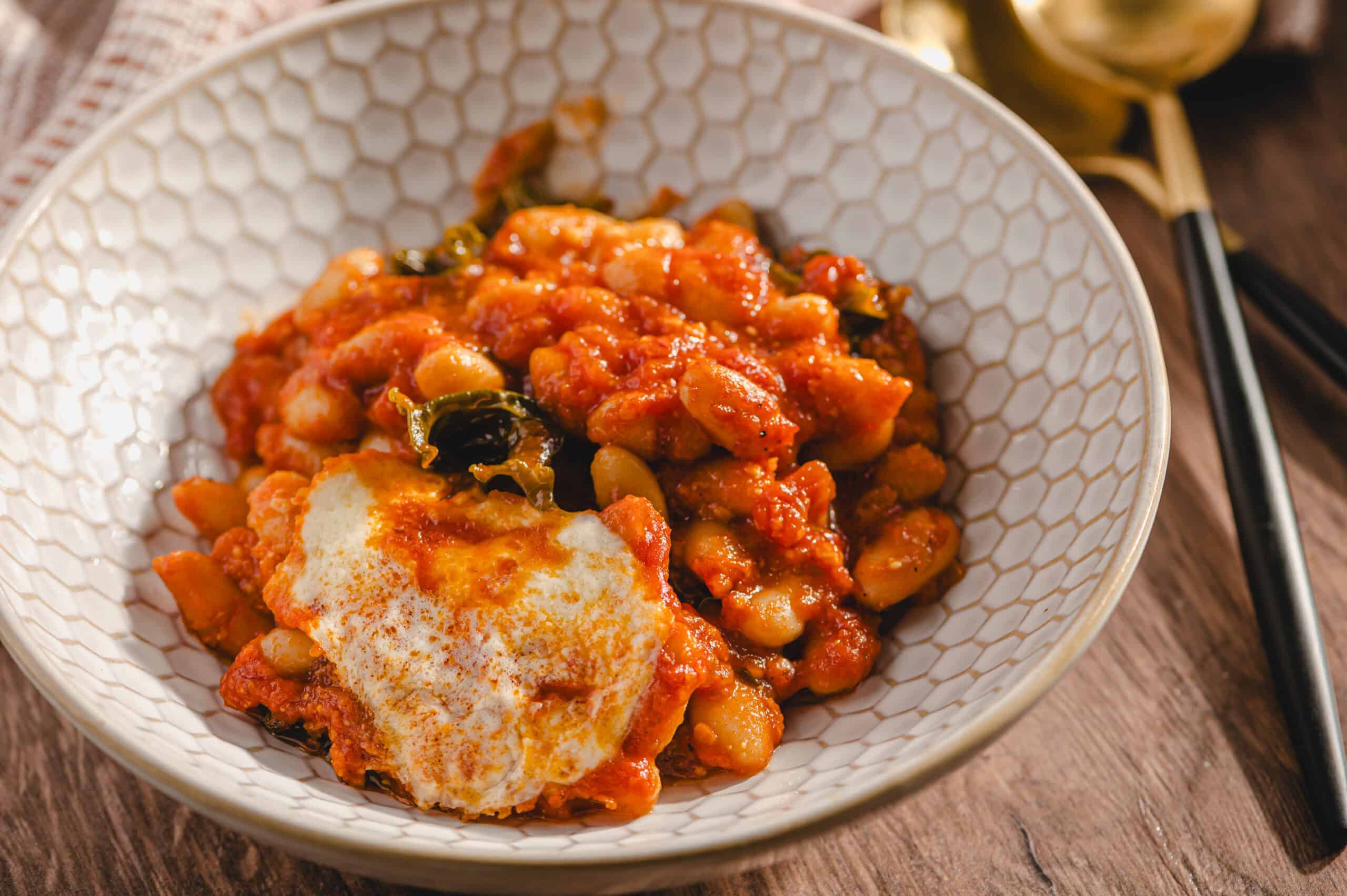 Savory bean and vegetable stew topped with melted cheese served in a textured white bowl, perfect for a hearty meal or comfort food.