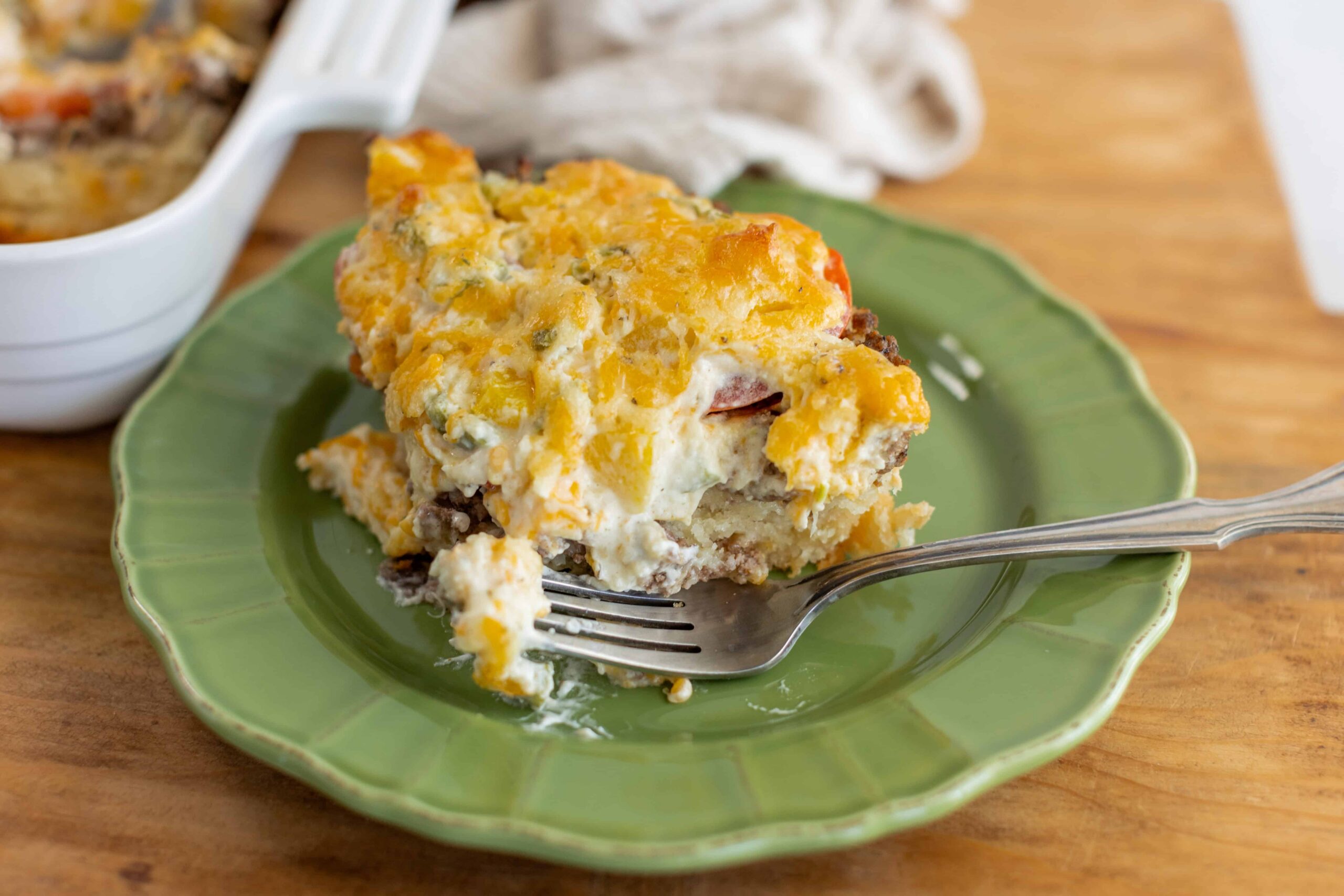 Cheesy breakfast casserole on a green plate with a fork, featuring melted cheese, eggs, and ground beef, served on a wooden table.