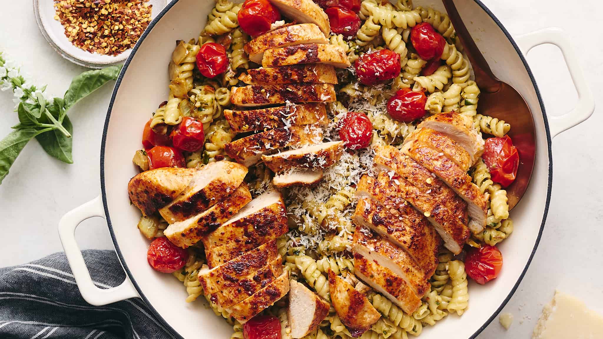 Grilled chicken pasta salad with cherry tomatoes, basil, and grated cheese in a white serving dish. Fresh ingredients and herbs are visible, creating an appetizing and colorful presentation.