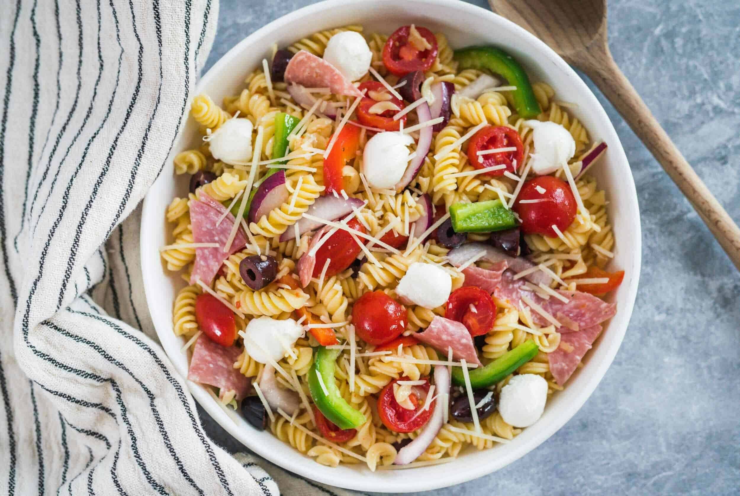 Colorful pasta salad with cherry tomatoes, mozzarella balls, olives, ham, and sliced onions, garnished with shredded cheese, served in a white bowl on a textured surface.