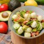Fresh cucumber and tomato salad with mozzarella balls, avocado, and herbs on a ceramic bowl. Styled with fresh ingredients like lemon, avocado, cucumber, and tomatoes on a rustic cloth background, perfect for healthy recipes.