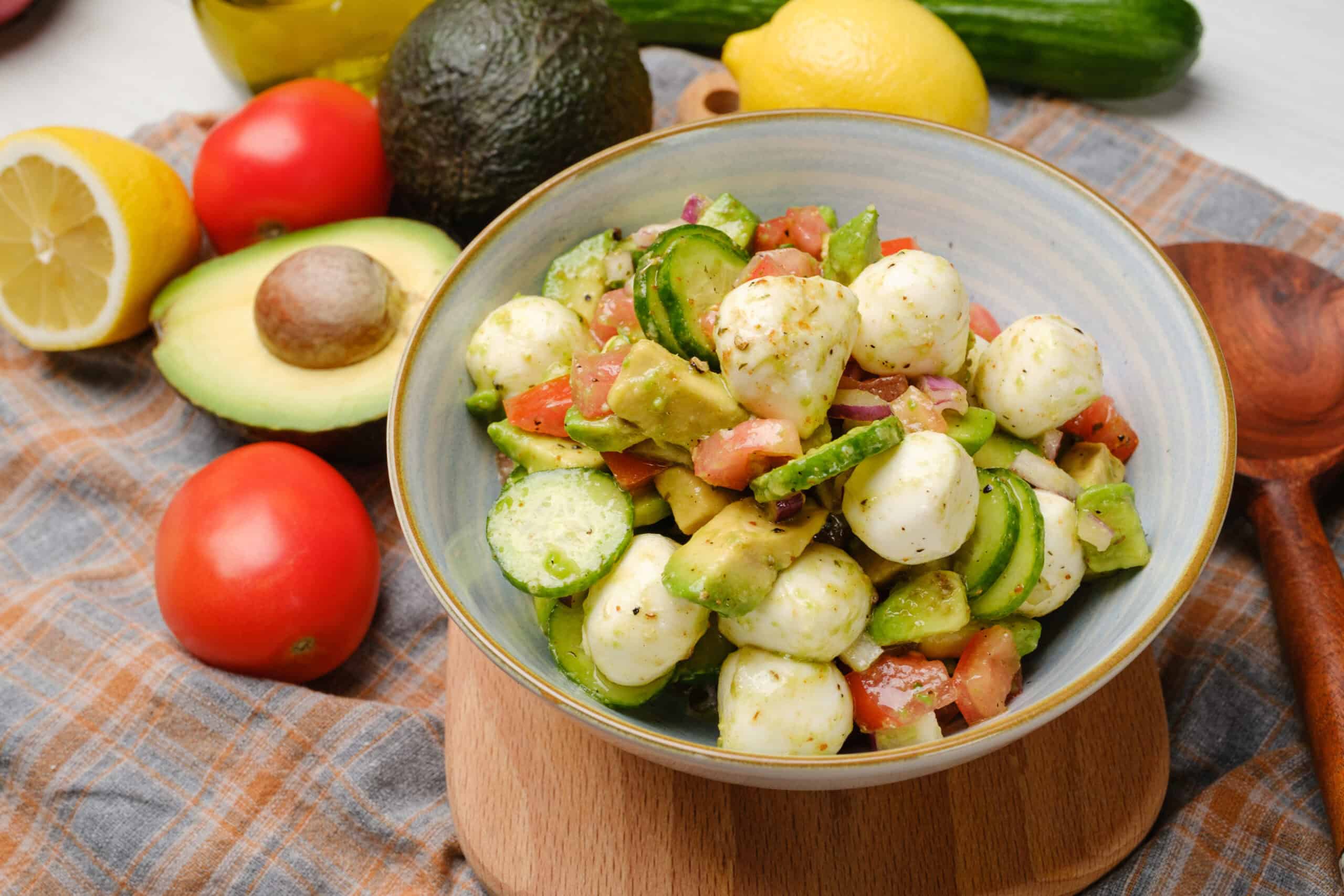Fresh cucumber and tomato salad with mozzarella balls, avocado, and herbs on a ceramic bowl. Styled with fresh ingredients like lemon, avocado, cucumber, and tomatoes on a rustic cloth background, perfect for healthy recipes.