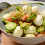 Fresh Italian Caprese Salad with mozzarella, avocado, cherry tomatoes, and cucumber, drizzled with olive oil and spices, served in a white bowl for a healthy, vegetarian appetizer or side dish.