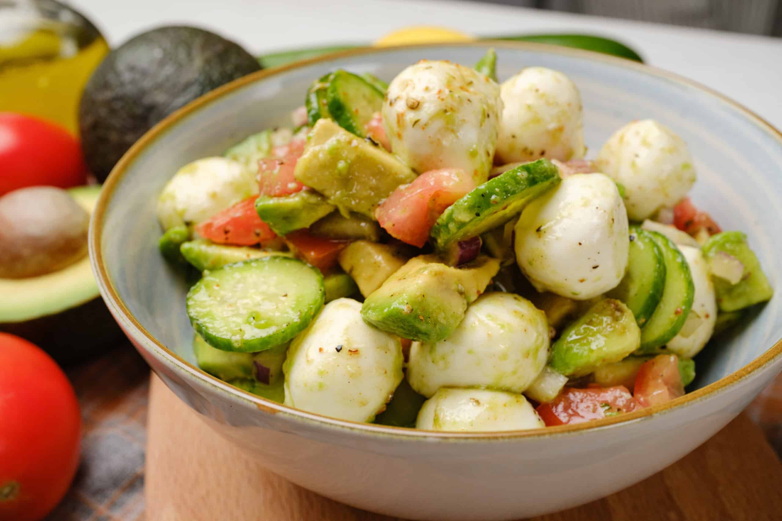 Fresh Italian Caprese Salad with mozzarella, avocado, cherry tomatoes, and cucumber, drizzled with olive oil and spices, served in a white bowl for a healthy, vegetarian appetizer or side dish.