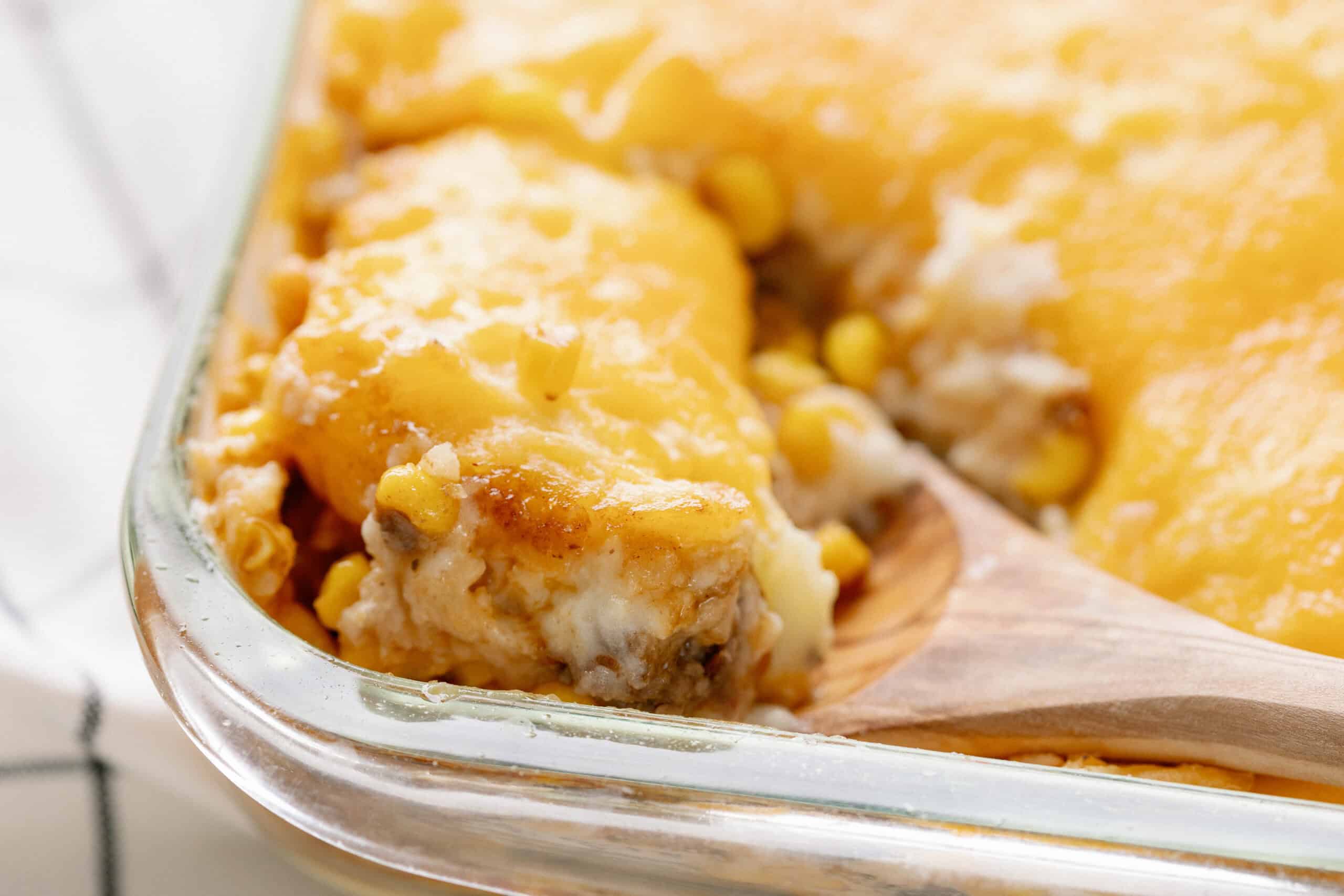 Creamy cheesy casserole with melted cheese, corn, and seasoned meat, served in a glass dish with a wooden spoon, perfect for comfort food recipes.
