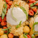 Crispy breaded chicken breasts topped with creamy burrata cheese, surrounded by roasted cherry tomatoes, fresh basil leaves, and caramelized yellow cherry tomatoes on a baking sheet.