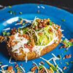 Crispy baked potato topped with guacamole, sour cream, shredded cheese, bacon bits, and chopped herbs on a blue plate.