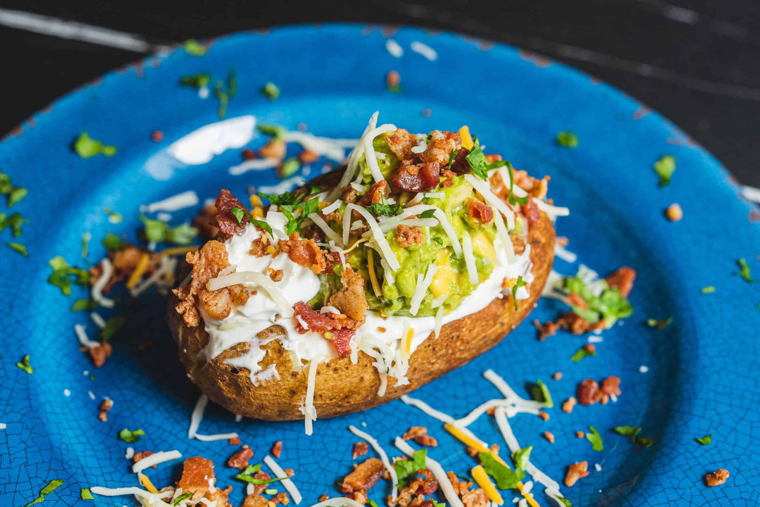 Crispy baked potato topped with guacamole, sour cream, shredded cheese, bacon bits, and chopped herbs on a blue plate.