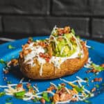 Soft baked potato topped with guacamole, sour cream, shredded cheese, bacon bits, and chopped green herbs on a vibrant blue plate.