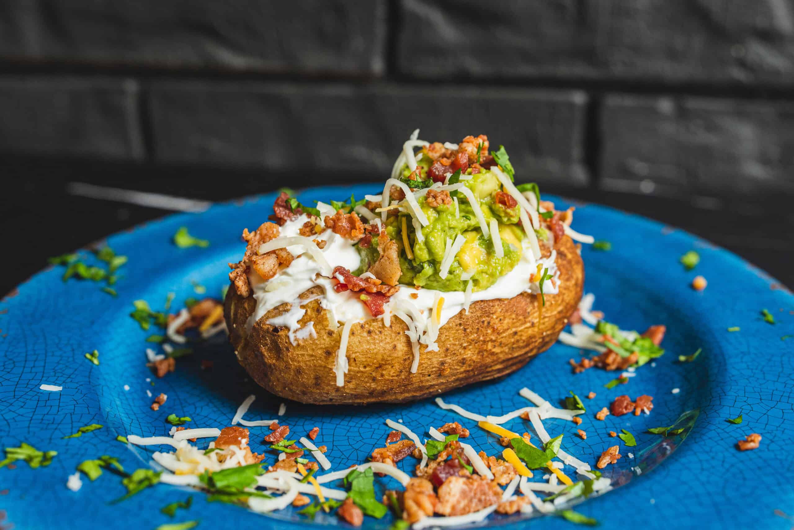 Soft baked potato topped with guacamole, sour cream, shredded cheese, bacon bits, and chopped green herbs on a vibrant blue plate.