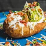 Creamy loaded baked potato topped with guacamole, shredded cheese, bacon bits, and sour cream on a blue plate. Perfect for a savory appetizer or comfort food meal.