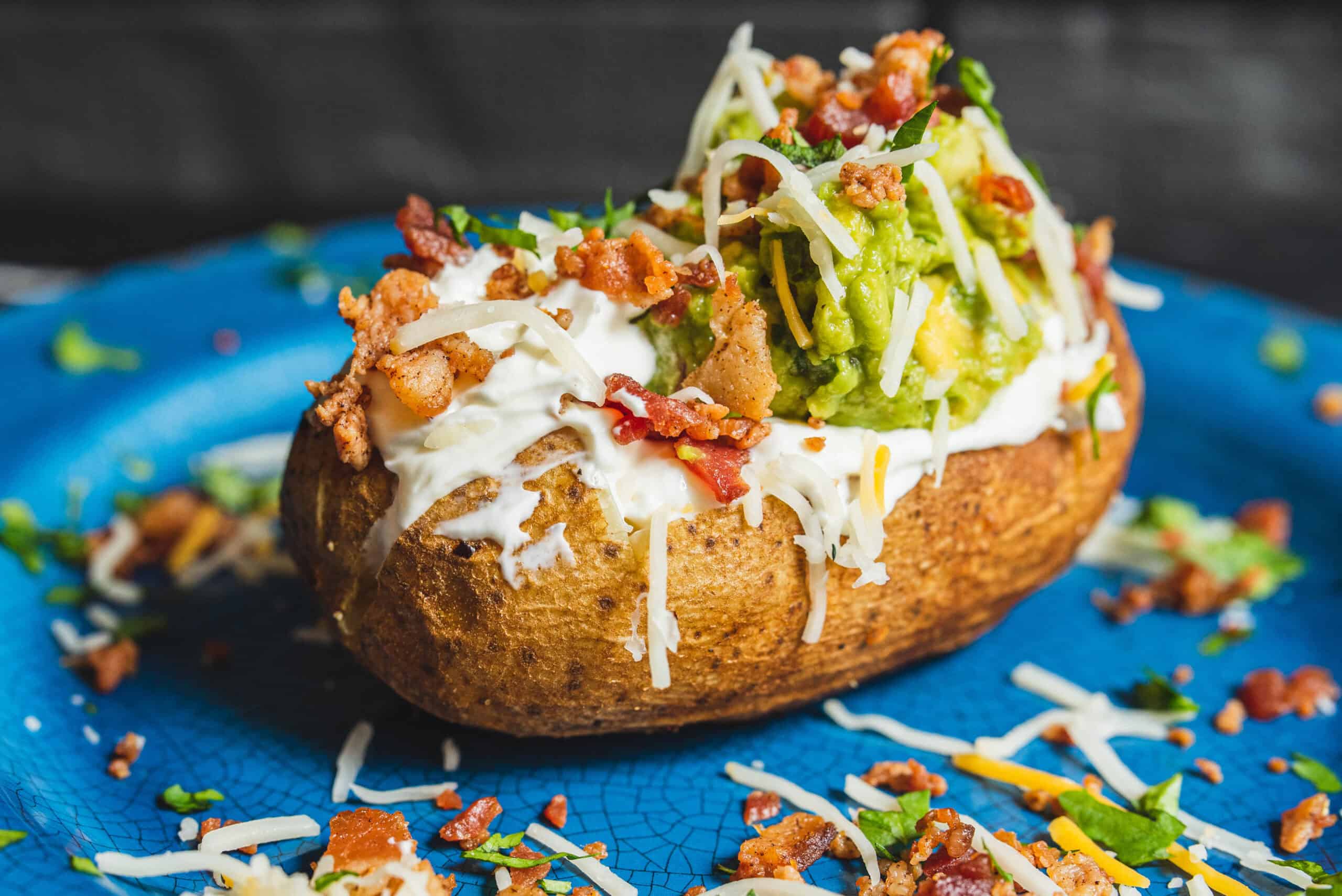 Creamy loaded baked potato topped with guacamole, shredded cheese, bacon bits, and sour cream on a blue plate. Perfect for a savory appetizer or comfort food meal.