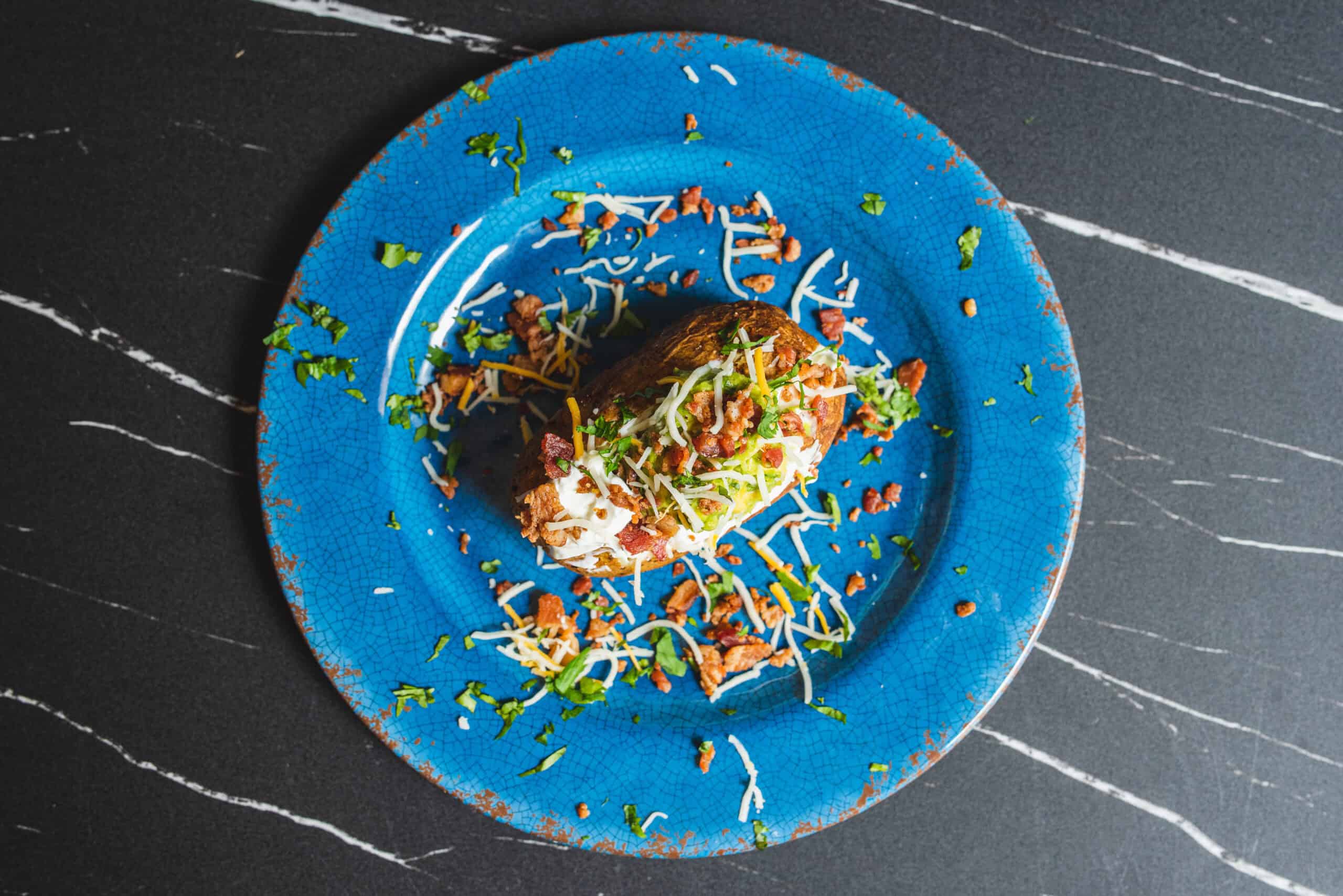 Savory loaded burrito on a vibrant blue plate, topped with shredded cheese, sour cream, bacon bits, chopped green onions, and fresh herbs, served on a dark textured background.