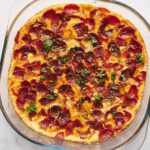 Golden crispy pepperoni pizza fresh out of the oven with melted cheese and herbs, served in a glass baking dish on a white marble surface.