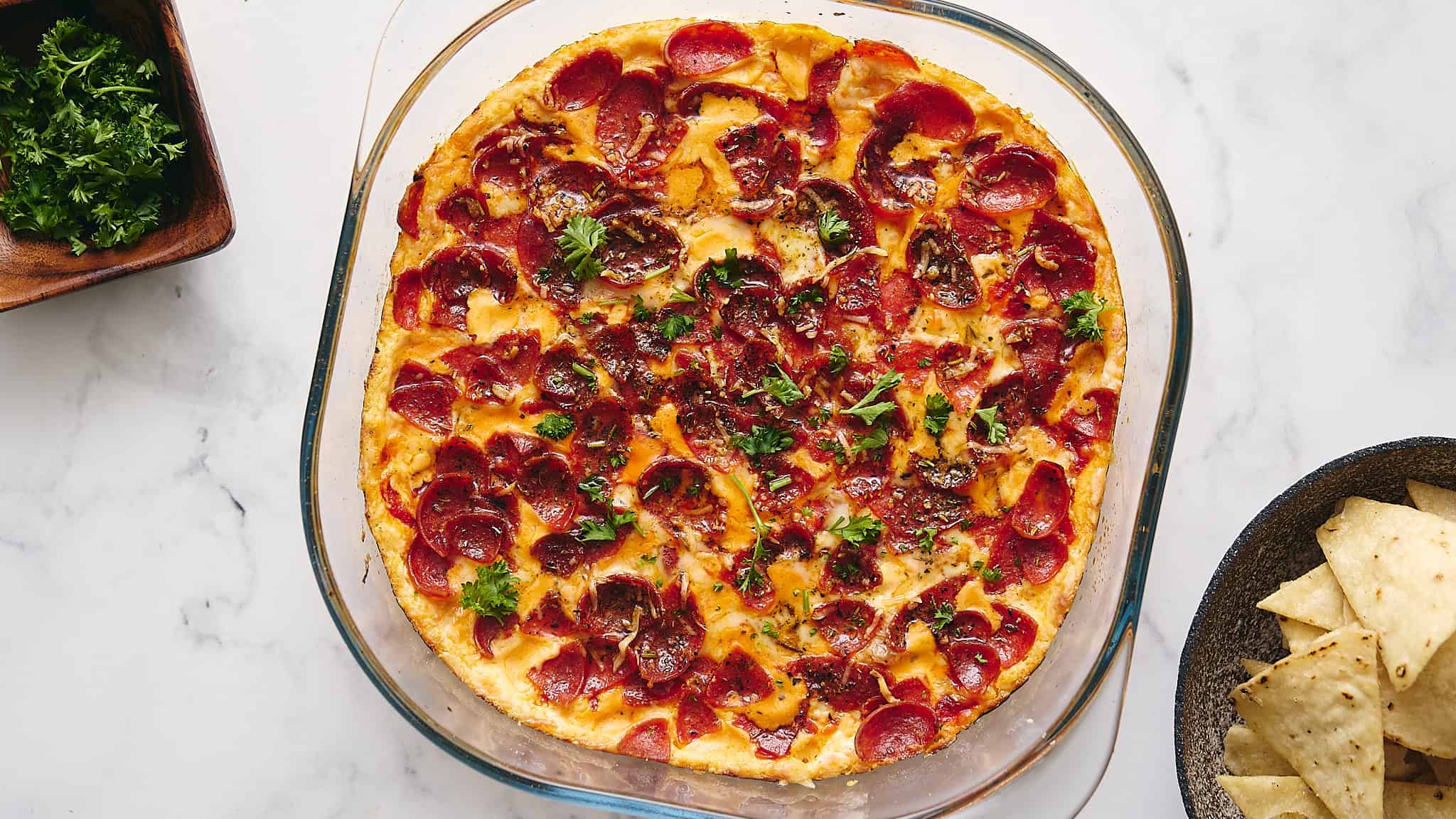 Golden crispy pepperoni pizza fresh out of the oven with melted cheese and herbs, served in a glass baking dish on a white marble surface.