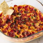 Juicy pepperoni pizza in a glass baking dish topped with fresh herbs, served with side tortilla chips and sliced tortilla chips in a skillet, on a white marble surface.