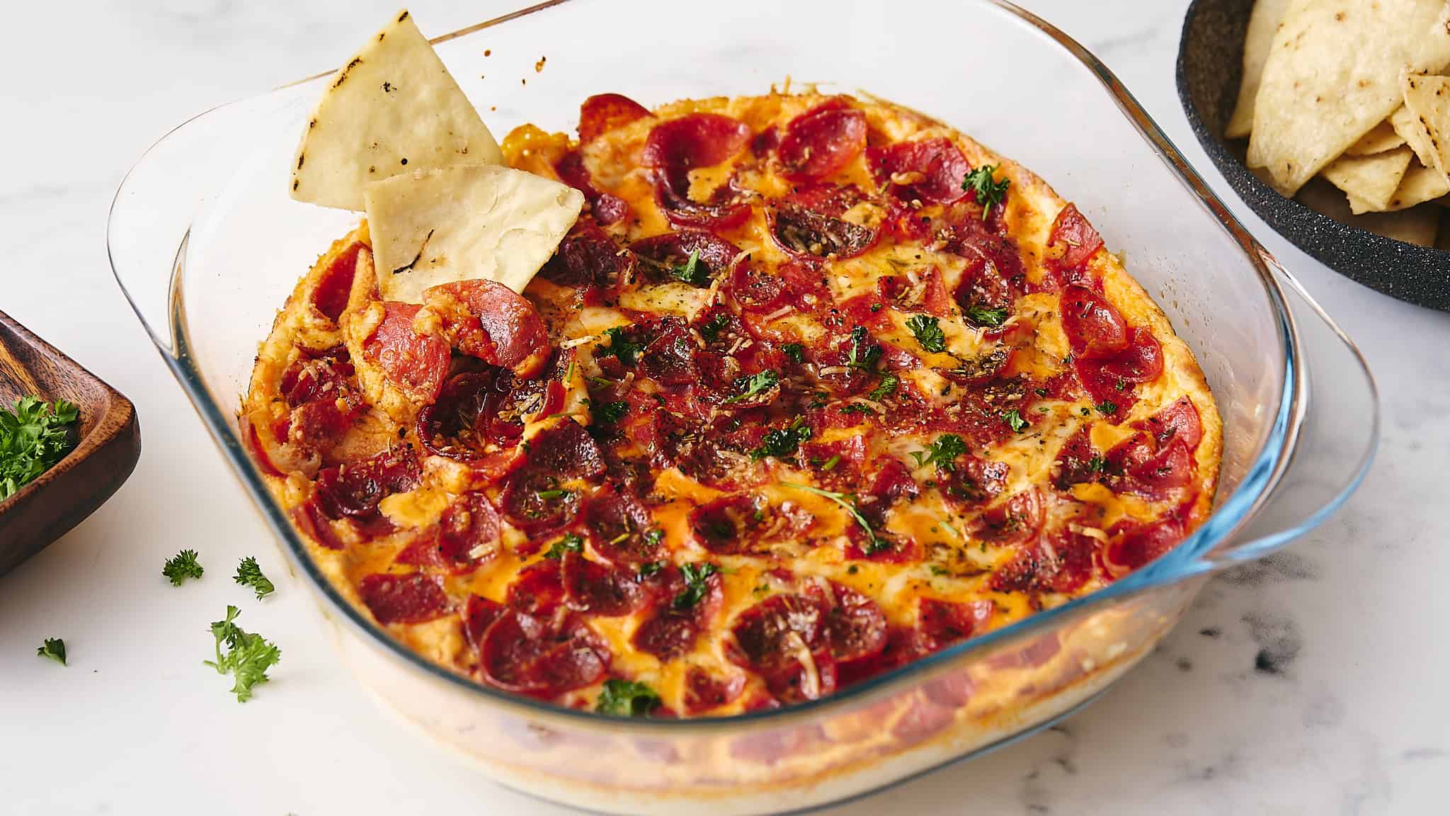 Juicy pepperoni pizza in a glass baking dish topped with fresh herbs, served with side tortilla chips and sliced tortilla chips in a skillet, on a white marble surface.