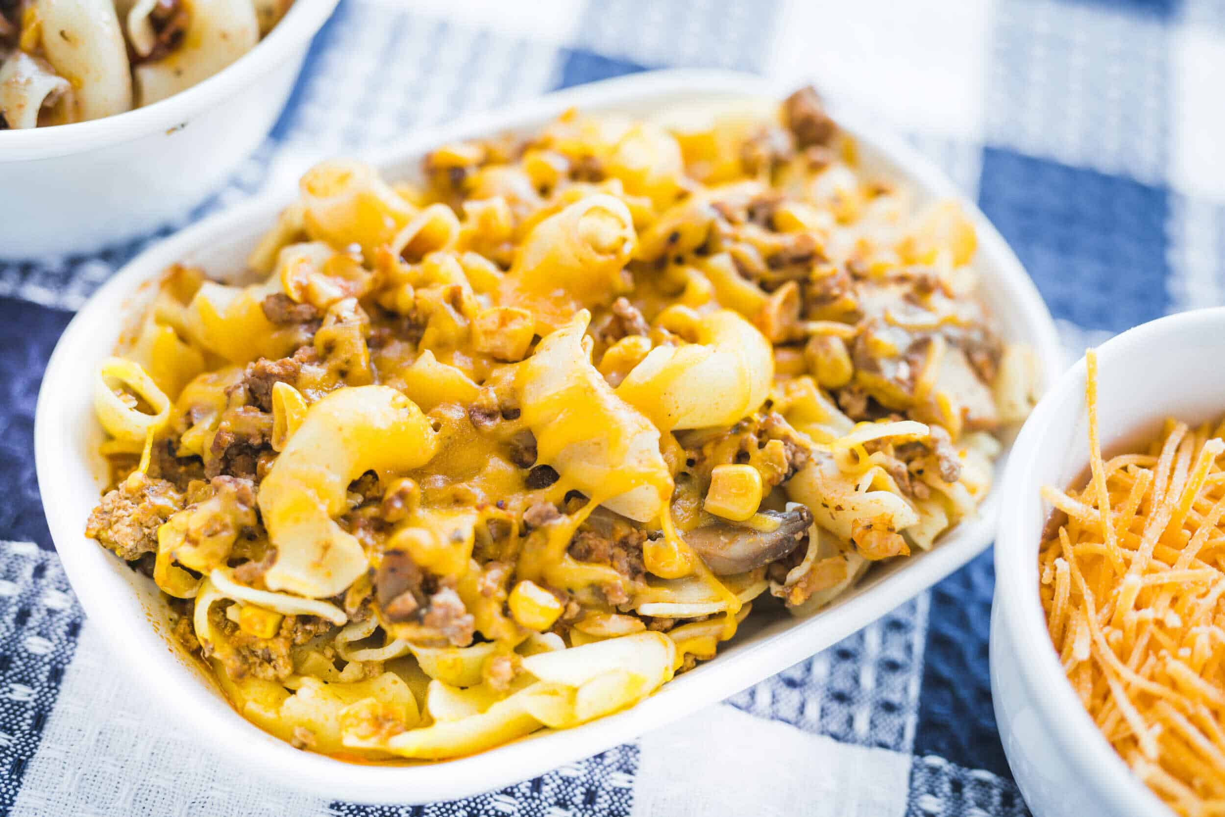 Creamy ground beef and pasta casserole topped with melted cheese, served in a white dish with side bowls of shredded cheese and chopped beef, perfect for comfort food lovers.