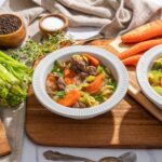 Creamy beef and vegetable noodle soup in white bowls, surrounded by fresh carrots, celery, herbs, spices, and cooking ingredients on a wooden cutting board.