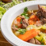 Savory beef vegetable soup with carrots, celery, onions, and herbs in a white bowl, served with fresh green vegetables on the side.