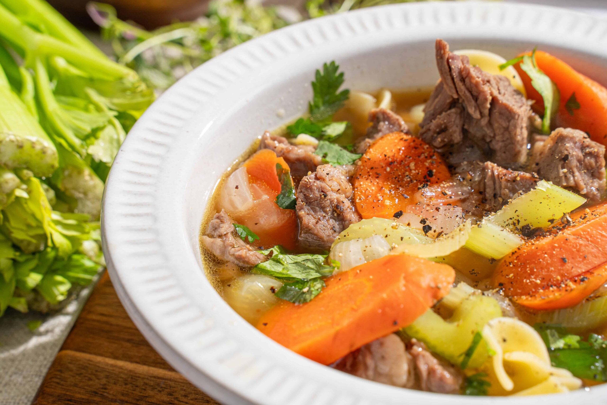 Savory beef vegetable soup with carrots, celery, onions, and herbs in a white bowl, served with fresh green vegetables on the side.