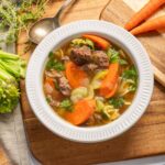 Hearty beef vegetable soup with carrots, celery, and fresh herbs served in a white bowl on a wooden surface.