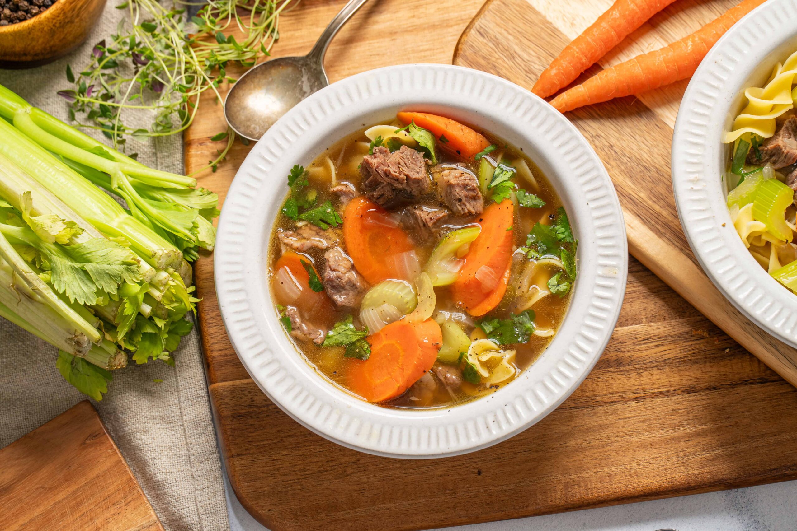 Hearty beef vegetable soup with carrots, celery, and fresh herbs served in a white bowl on a wooden surface.