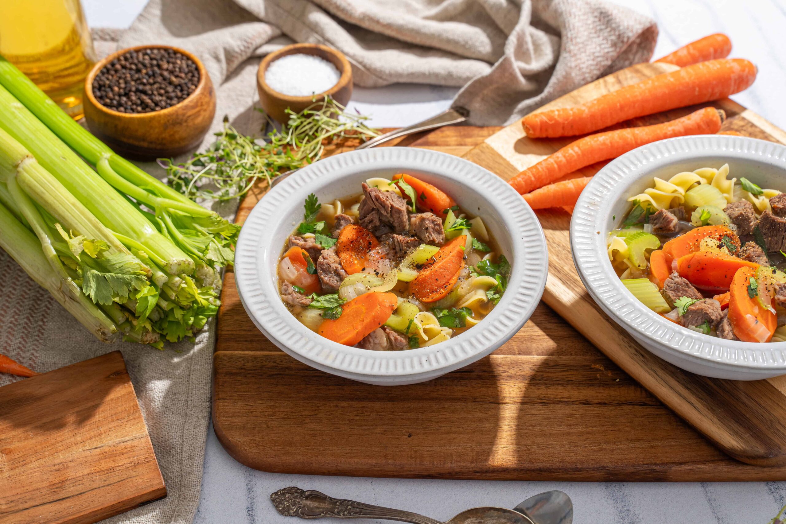 Creamy beef and vegetable noodle soup in white bowls, surrounded by fresh carrots, celery, herbs, spices, and cooking ingredients on a wooden cutting board.