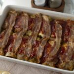 Slices of bacon layered over a casserole dish with vegetables and tomato sauce, ready for baking.