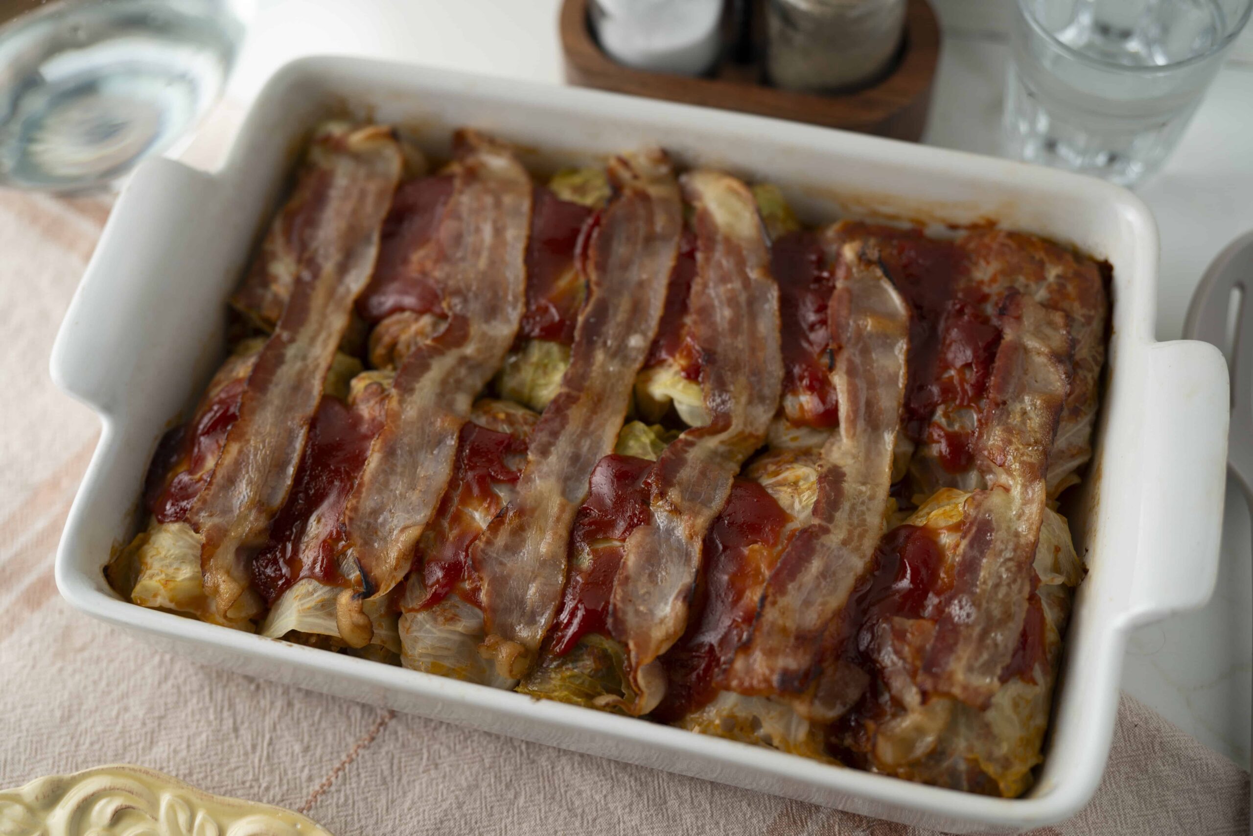Slices of bacon layered over a casserole dish with vegetables and tomato sauce, ready for baking.