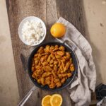 Crispy orange chicken stir-fry served in a black skillet with chopped carrots, accompanied by a bowl of steamed white rice, fresh lemon wedges, and seasonings on a rustic wooden table for a flavorful meal.