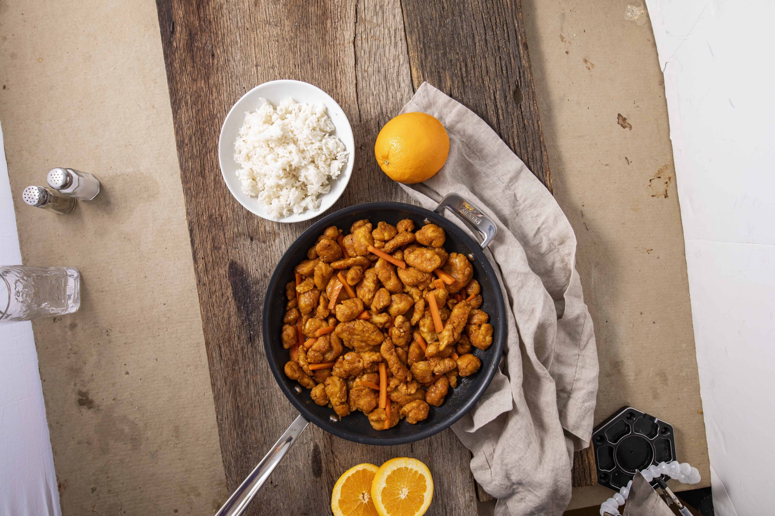 Crispy orange chicken stir-fry served in a black skillet with chopped carrots, accompanied by a bowl of steamed white rice, fresh lemon wedges, and seasonings on a rustic wooden table for a flavorful meal.