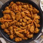 Crispy orange chicken stir-fry in a black skillet with carrots, served with white rice, fresh orange slices, and a whole orange on a rustic wooden table.