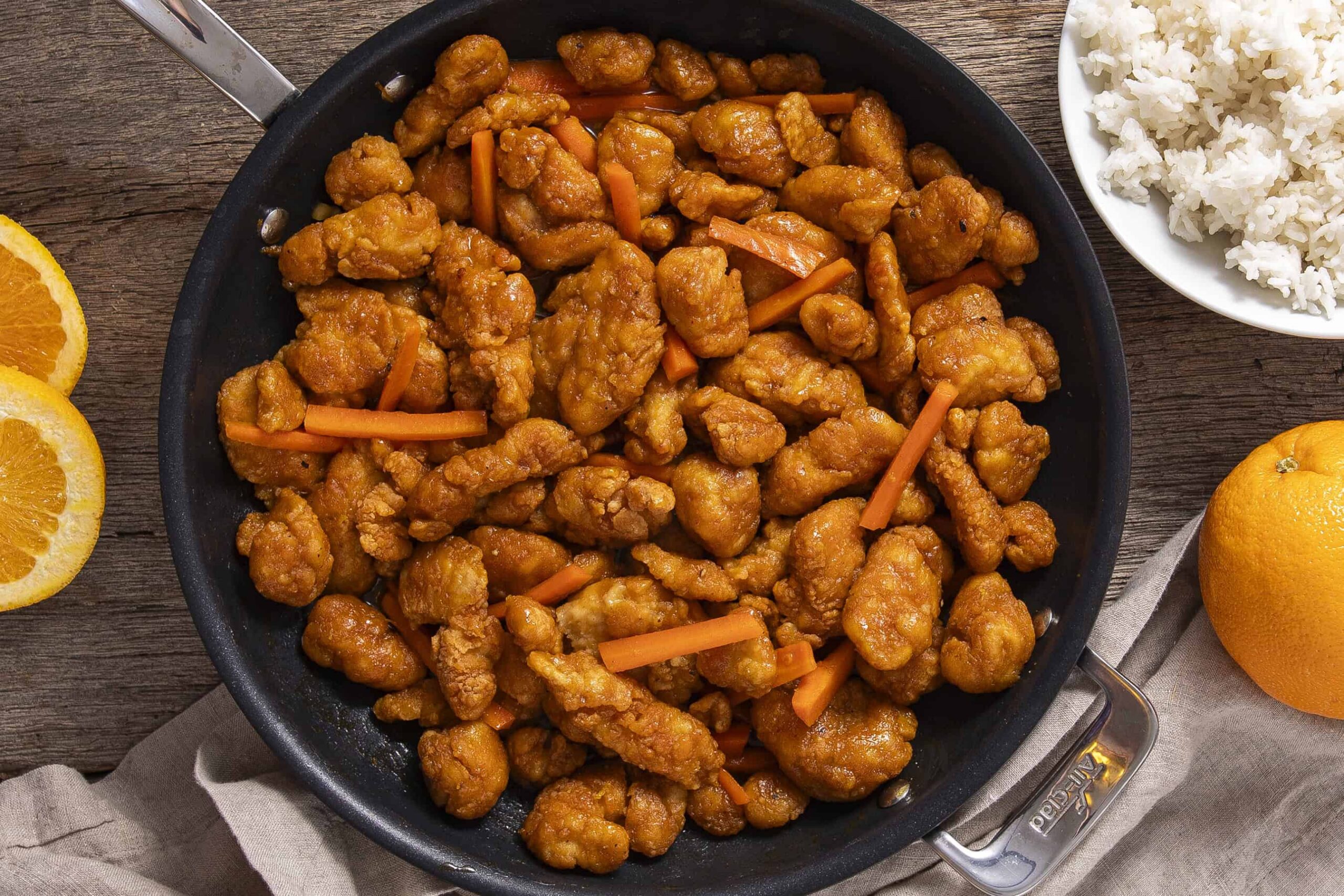 Crispy orange chicken stir-fry in a black skillet with carrots, served with white rice, fresh orange slices, and a whole orange on a rustic wooden table.