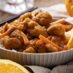 Crispy orange chicken served over white rice with lemon wedges and shredded carrots in a white bowl, featuring a flavorful Asian-inspired dish with vibrant orange sauce.