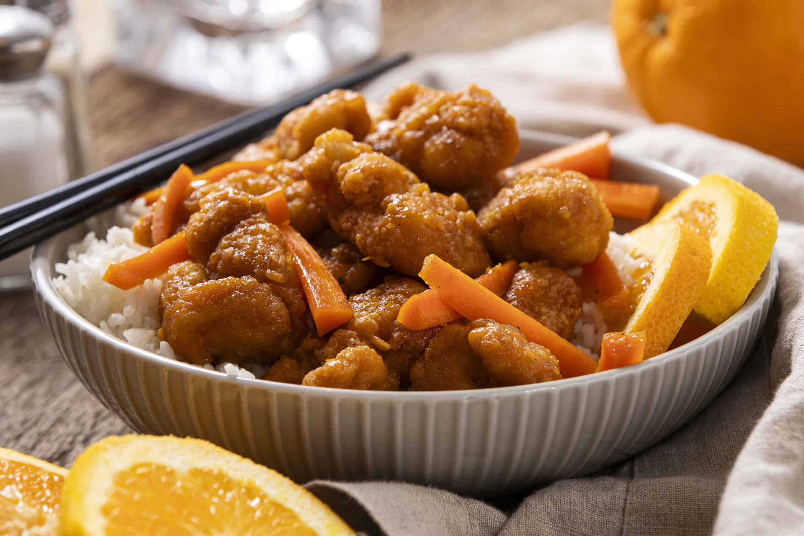 Crispy orange chicken served over white rice with lemon wedges and shredded carrots in a white bowl, featuring a flavorful Asian-inspired dish with vibrant orange sauce.