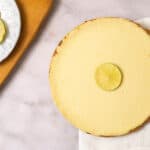 Lemon cheesecake topped with a lemon slice on a white plate, featuring a graham cracker crust, served on a white cloth with fresh lemon slices nearby. Perfect for summer desserts.