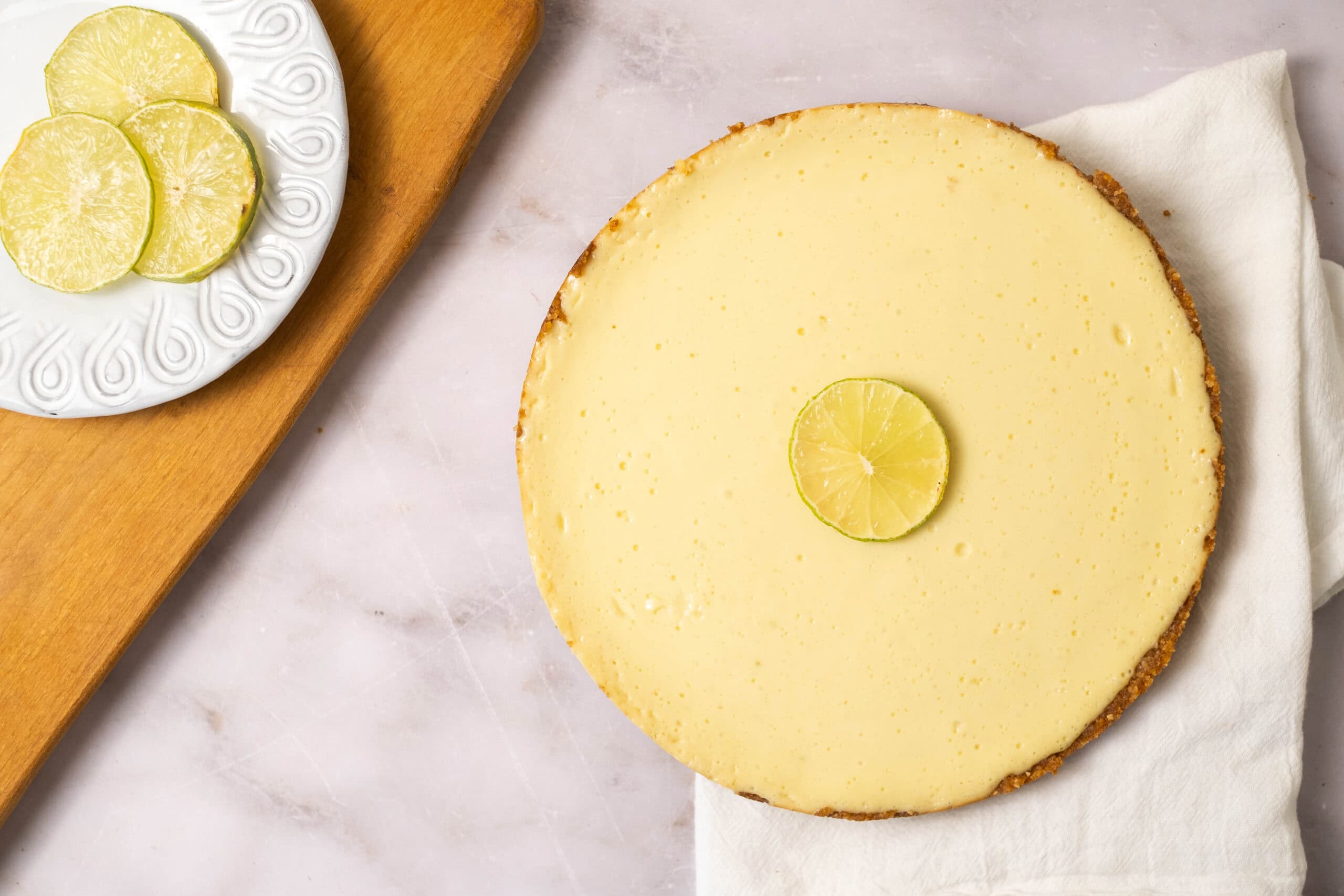 Lemon cheesecake topped with a lemon slice on a white plate, featuring a graham cracker crust, served on a white cloth with fresh lemon slices nearby. Perfect for summer desserts.
