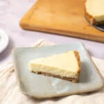 Creamy cheesecake slice on a light green plate, with a graham cracker crust and smooth, rich topping, served with fresh lemon slices in the background. Perfect dessert for cheesecake lovers and gatherings.