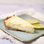 Creamy lemon cheesecake with graham cracker crust served on a light blue plate, garnished with fresh lime slices. Perfect for a refreshing dessert.