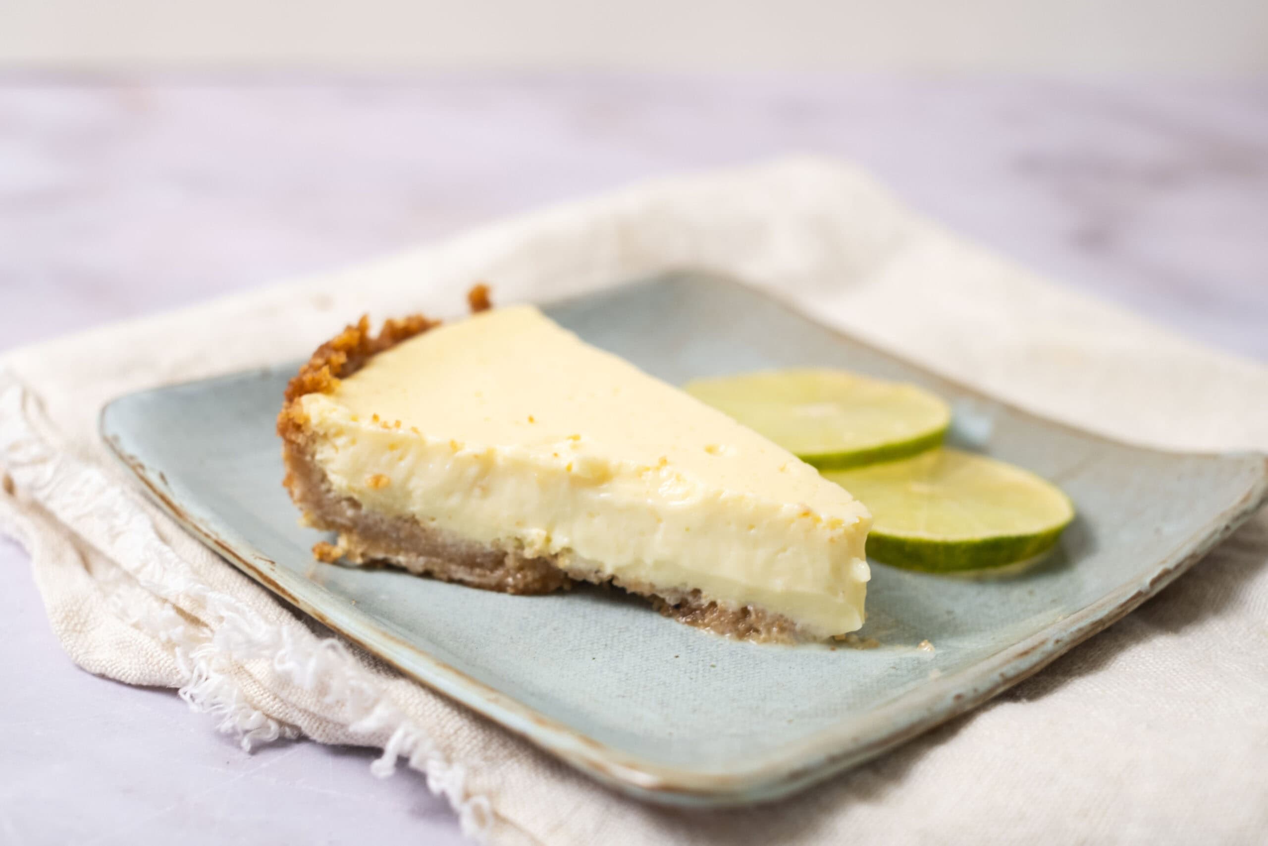 Creamy lemon cheesecake with graham cracker crust served on a light blue plate, garnished with fresh lime slices. Perfect for a refreshing dessert.