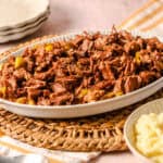 Slow cooked shredded beef in a white serving dish with chopped pickles, served on a woven placemat, accompanied by mashed potatoes and a stack of plates, perfect comfort food for family dinner.