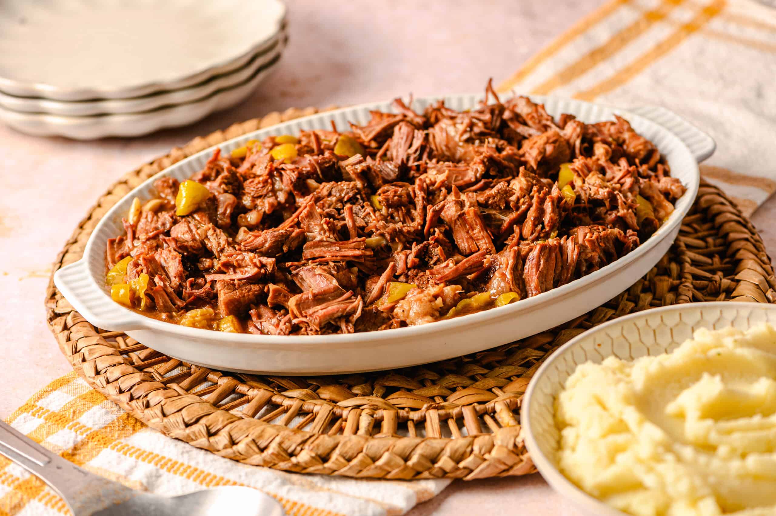Slow cooked shredded beef in a white serving dish with chopped pickles, served on a woven placemat, accompanied by mashed potatoes and a stack of plates, perfect comfort food for family dinner.