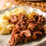 Slow cooker shredded beef with mashed potatoes on a white decorative plate, served with green pickled peppers, ready for a hearty meal.